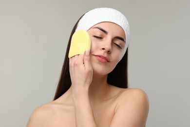 Young woman with headband washing her face using sponge on light grey background