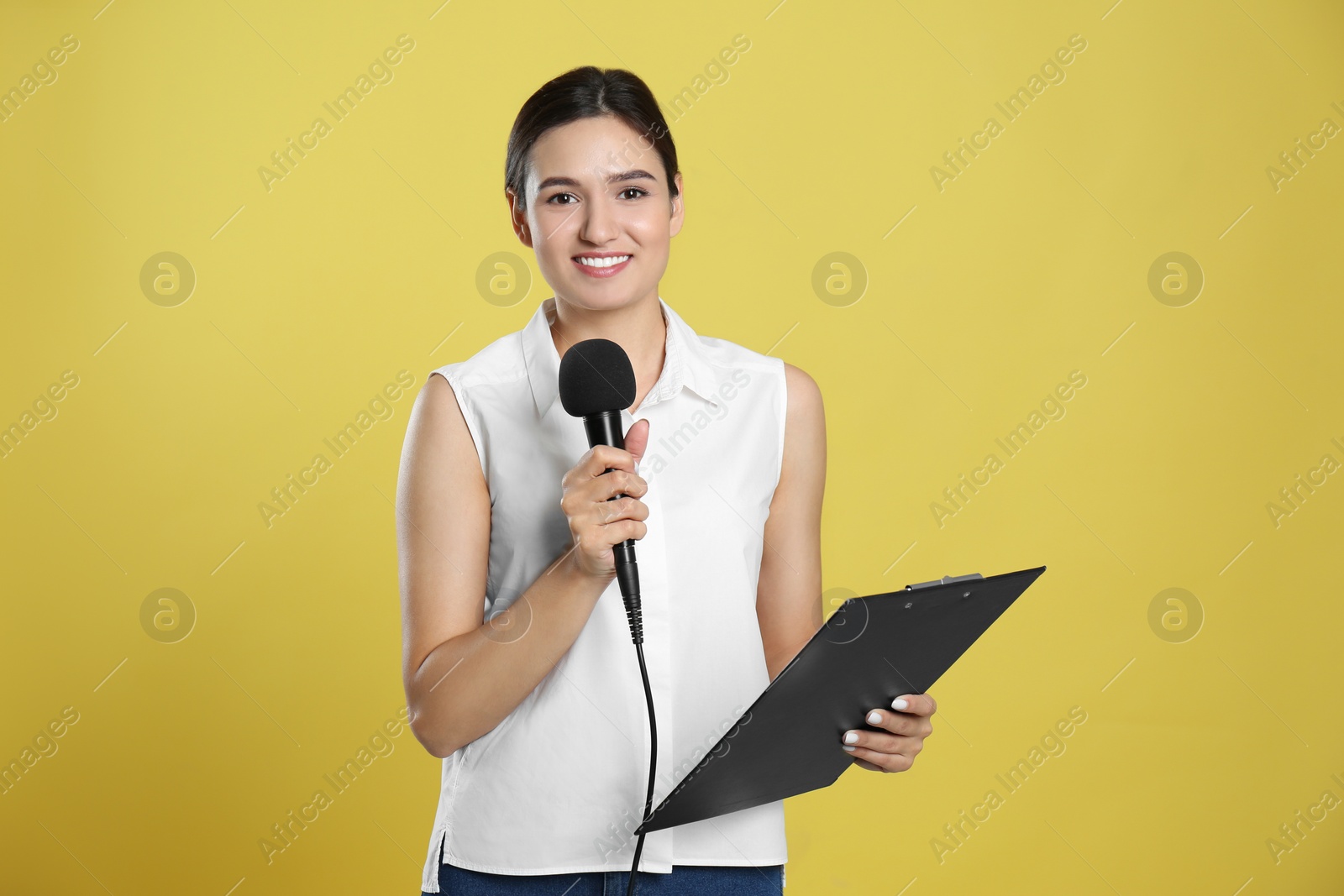 Photo of Young female journalist with microphone and clipboard on yellow background