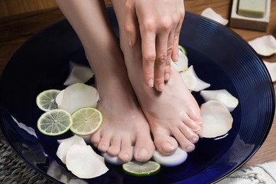 Photo of Woman soaking her feet in bowl with water, petals and lime slices on floor, closeup. Spa treatment