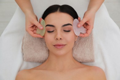 Photo of Young woman receiving facial massage with gua sha tools in beauty salon, top view