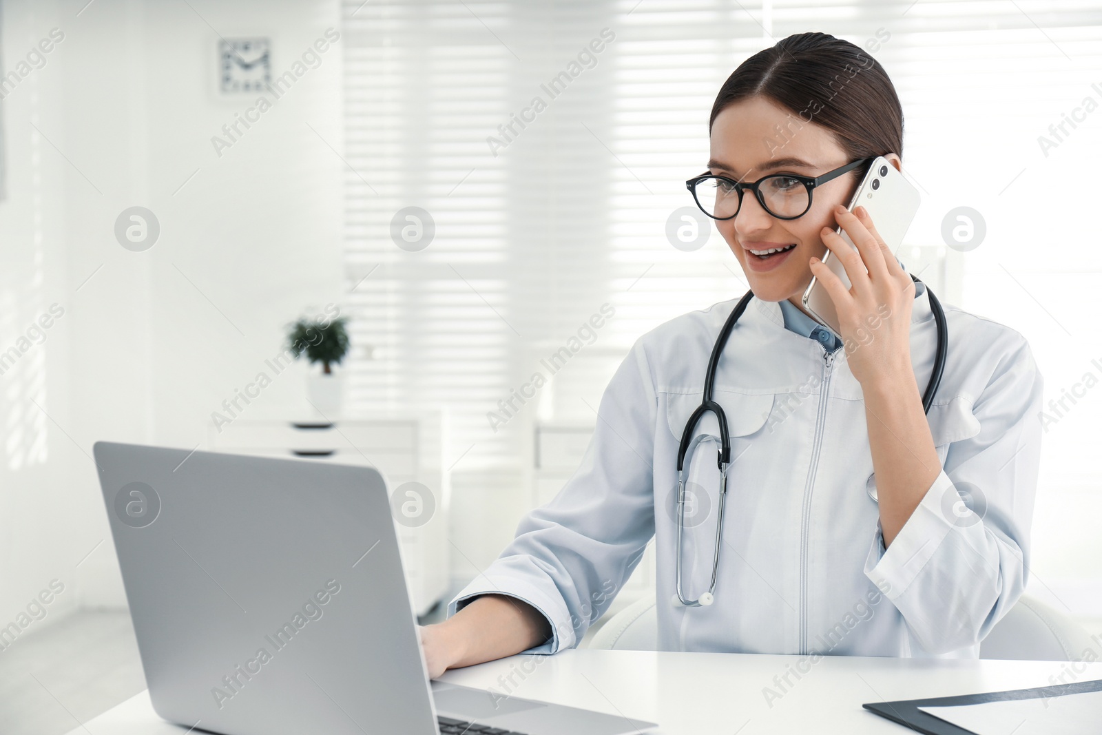 Photo of Young female doctor talking on phone at table in office