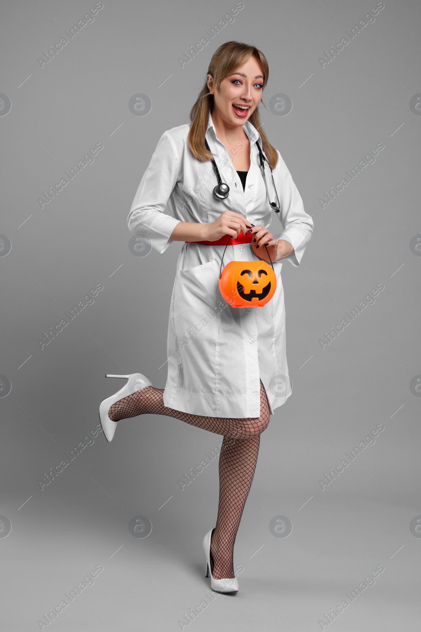 Photo of Happy woman in scary nurse costume with pumpkin bucket on light grey background. Halloween celebration