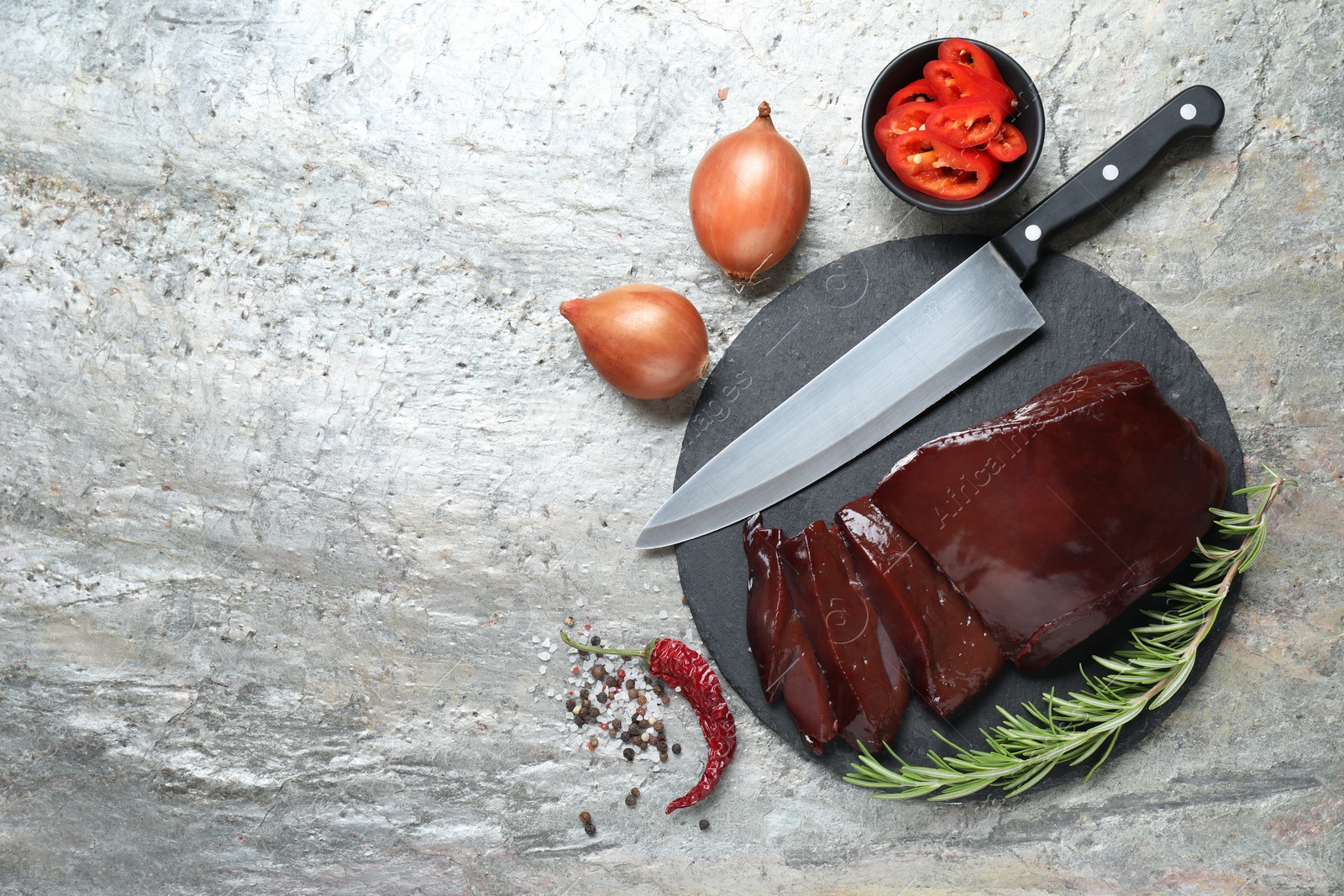 Photo of Cut raw beef liver with rosemary, chili pepper, onions and knife on grey table, flat lay. Space for text