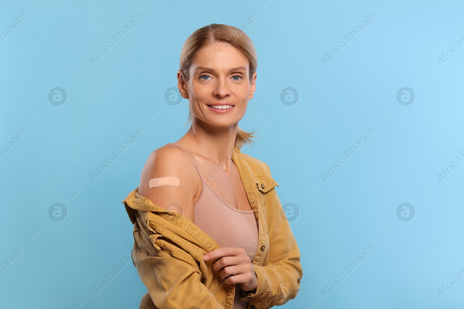 Photo of Smiling woman with adhesive bandage on arm after vaccination on light blue background