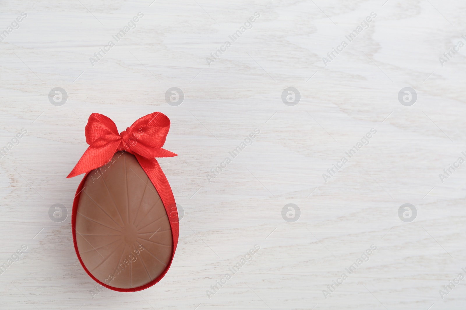 Photo of Sweet chocolate egg with red bow on white wooden table, top view. Space for text