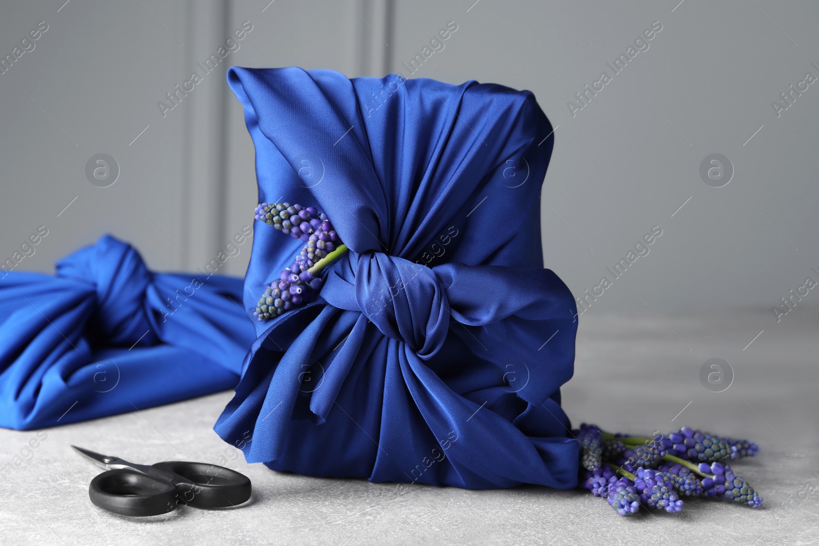 Photo of Furoshiki technique. Gifts packed in blue silk fabric, muscari flowers, ribbon and scissors on light grey table