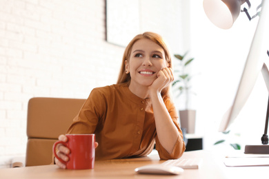 Young woman with cup of drink relaxing at table in office during break