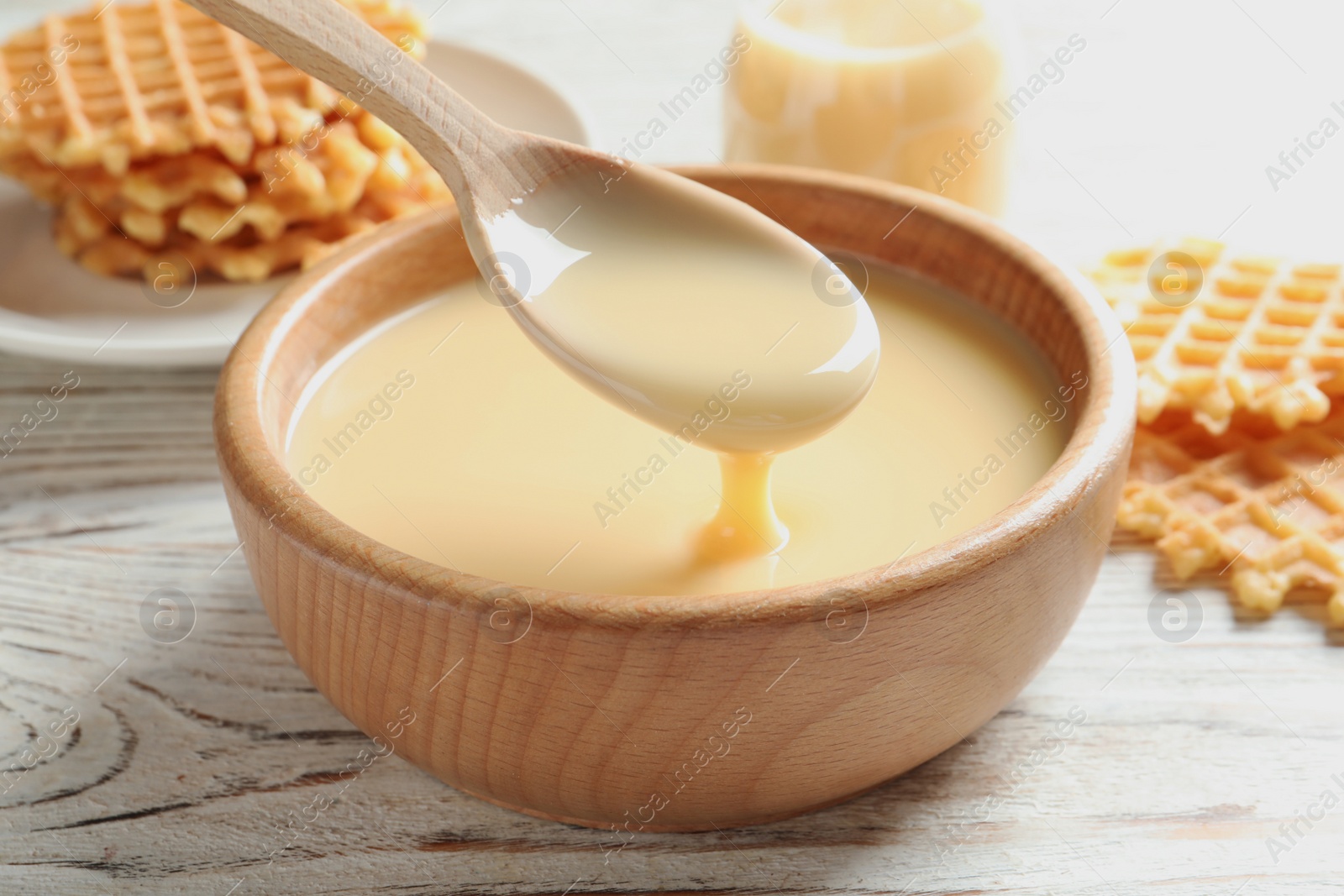 Photo of Spoon of pouring condensed milk over bowl on table, closeup. Dairy products