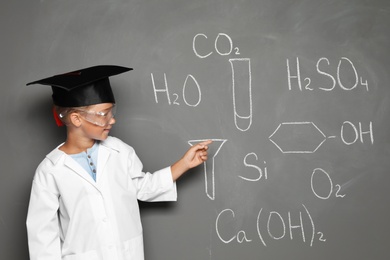 Little school child in laboratory uniform with graduate cap and chemical formulas on grey background