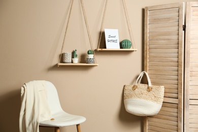 Comfortable chair and wooden shelves with different decorative elements on beige wall in room interior