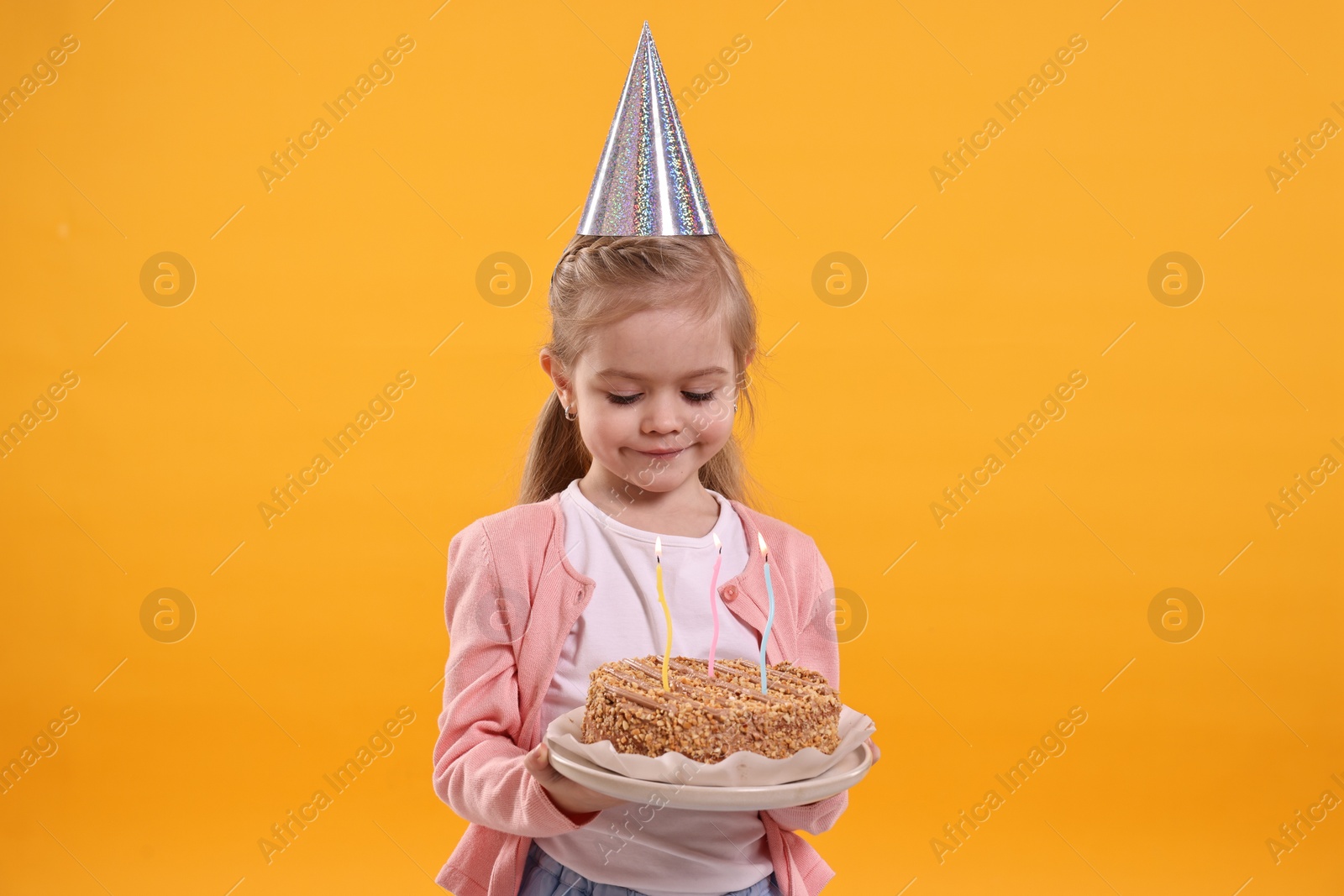 Photo of Birthday celebration. Cute little girl in party hat holding tasty cake with burning candles on orange background