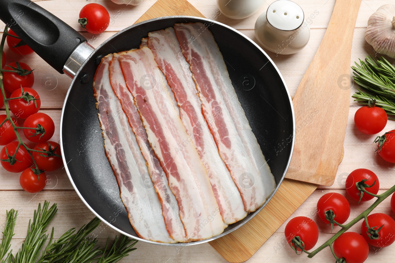 Photo of Slices of raw bacon in frying pan and fresh products on wooden table, flat lay