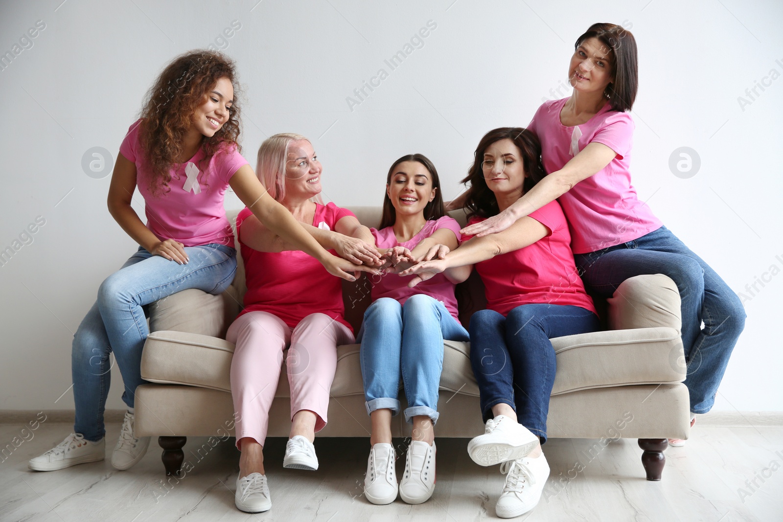 Photo of Group of women with silk ribbons joining hands on sofa against light wall. Breast cancer awareness concept