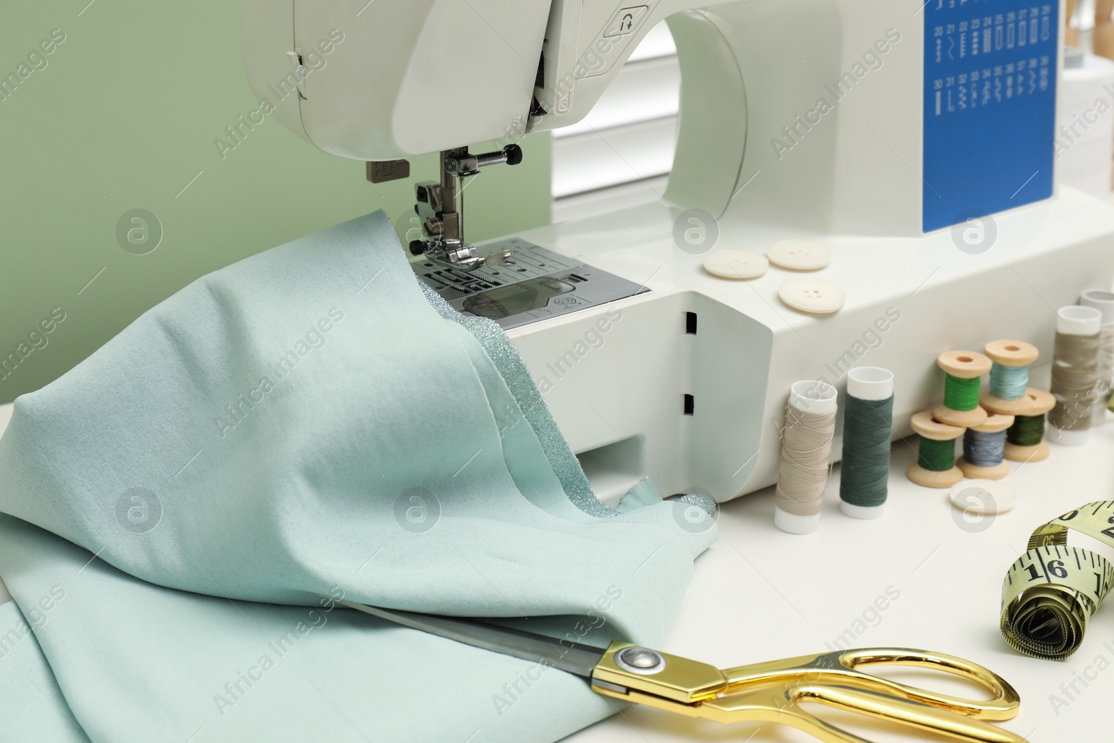 Photo of Sewing machine with fabric, scissors, measuring tape and threads on white table, closeup