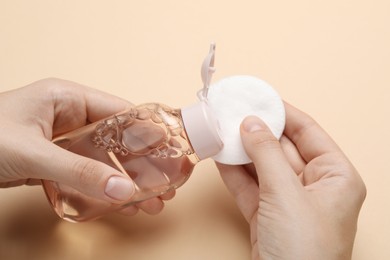 Photo of Woman pouring micellar water from bottle onto cotton pad against beige background, closeup
