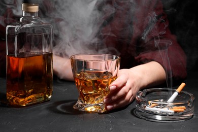 Photo of Alcohol addiction. Woman with whiskey and smoldering cigarettes at dark textured table, closeup