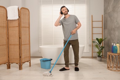 Photo of Enjoying cleaning. Man in headphones listening to music and mopping floor in bathroom