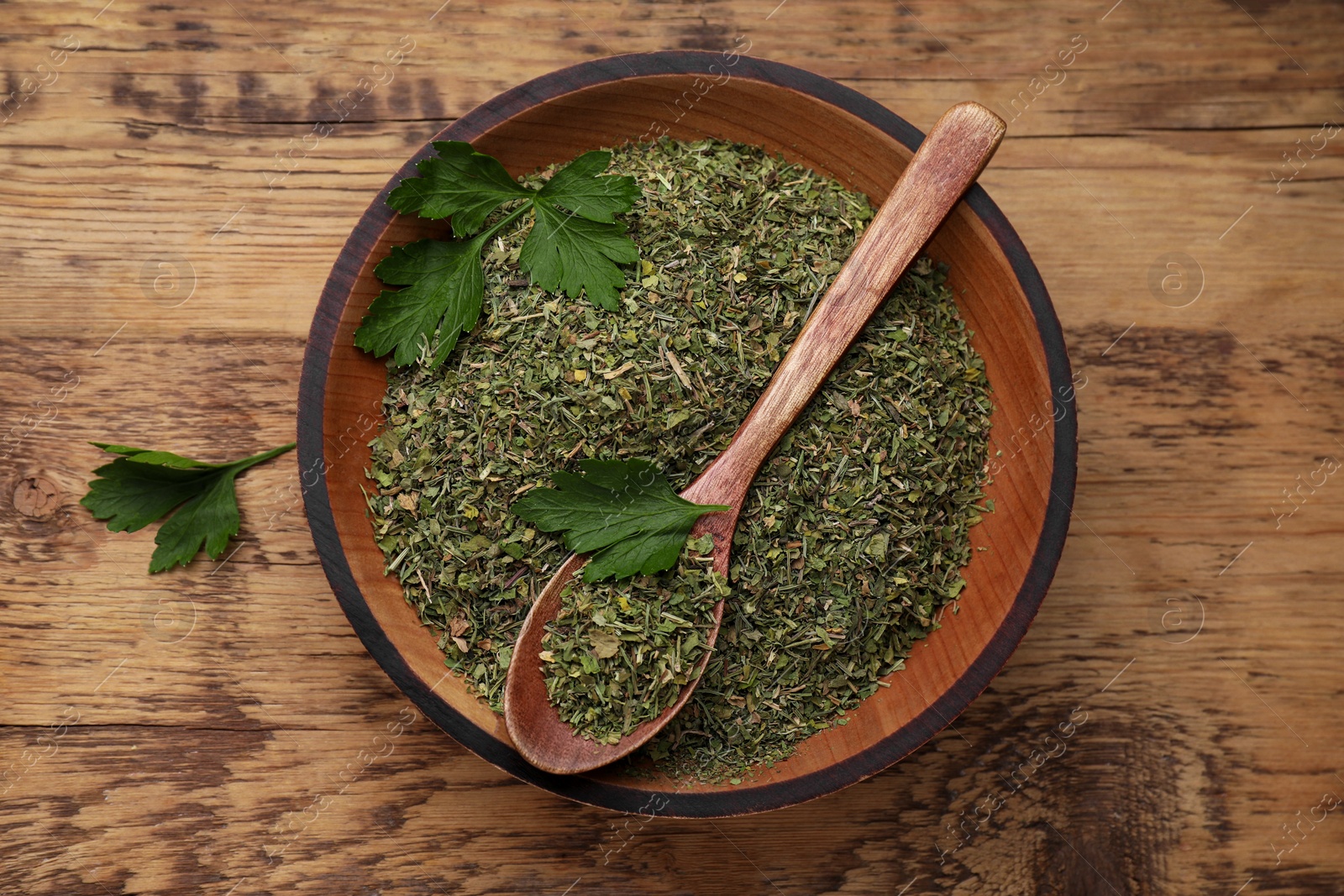 Photo of Bowl and spoon with dried parsley on wooden table, top view