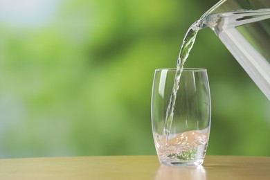 Photo of Pouring fresh water from jug into glass at wooden table against blurred green background, closeup. Space for text