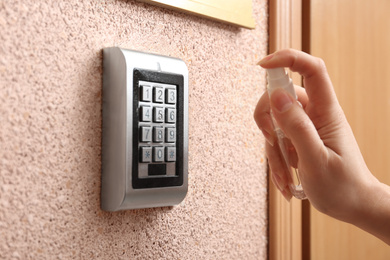 Photo of Woman spraying antiseptic onto electronic lock's keypad indoors, closeup