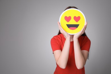 Woman covering face with heart eyes emoji on grey background, space for text