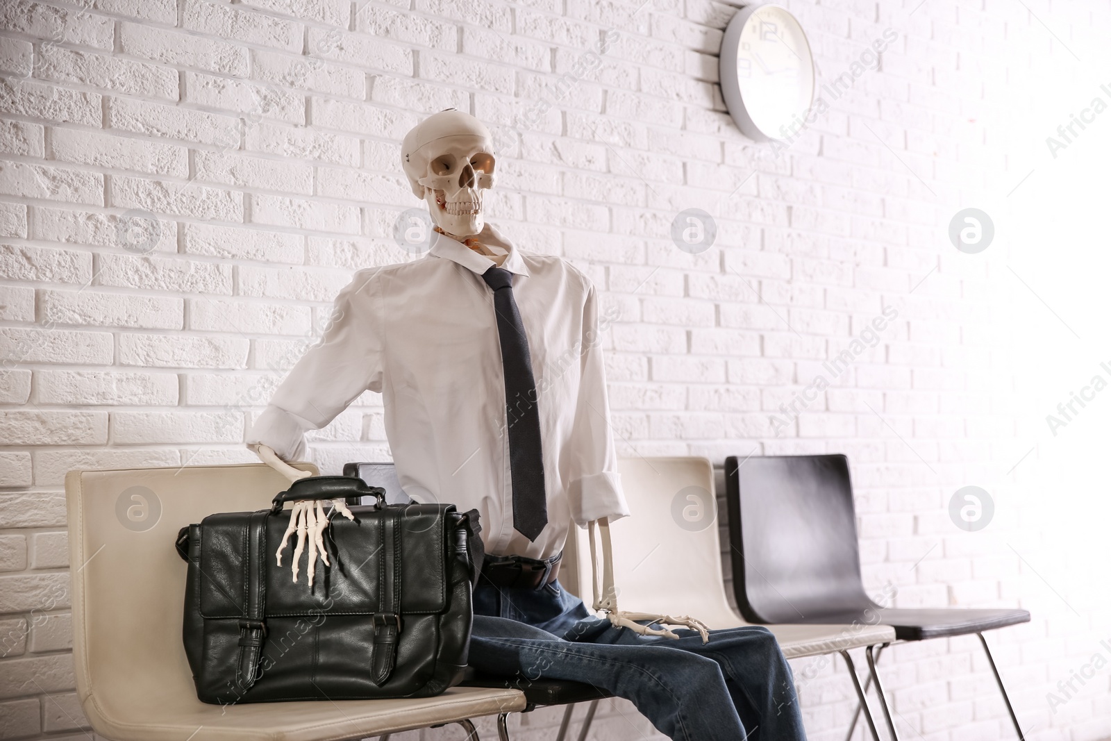 Photo of Human skeleton in office wear sitting on chair near brick wall indoors