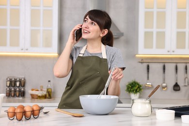 Happy young housewife talking on smartphone while cooking at white marble table in kitchen