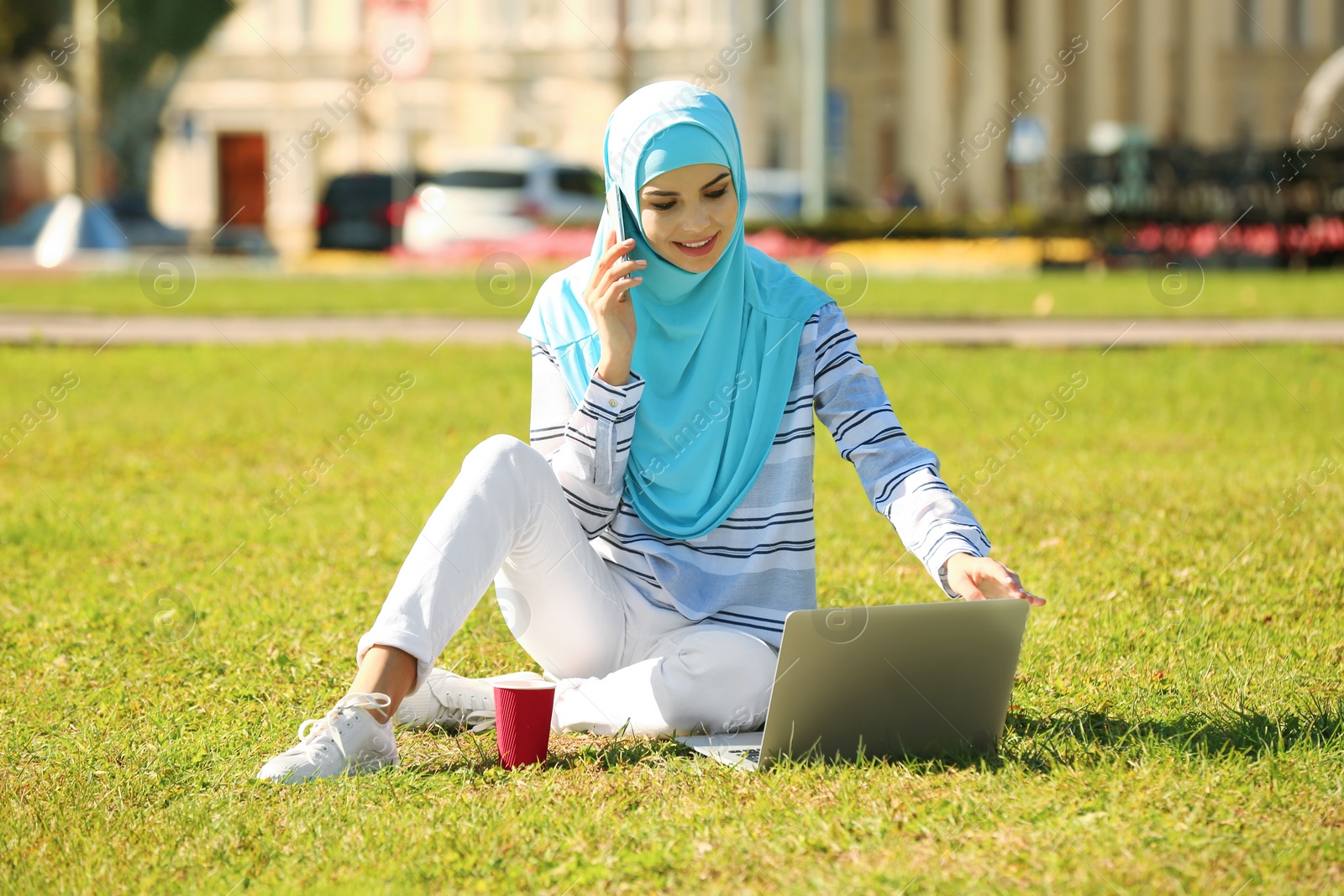 Photo of Muslim woman in hijab with laptop talking on phone outdoors