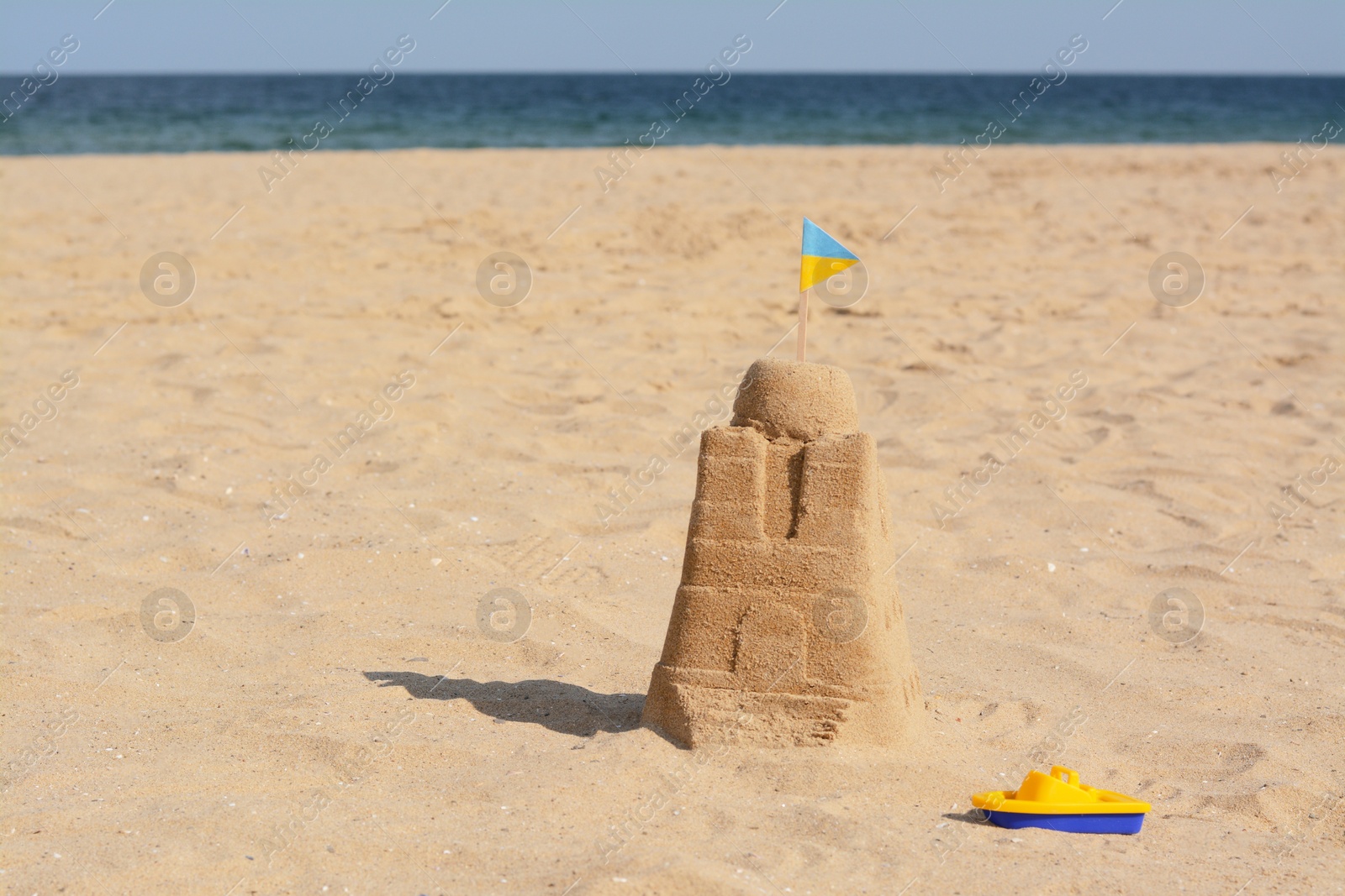 Photo of Beautiful sand castle with small Ukrainian flag and toy boat on beach near sea, space for text