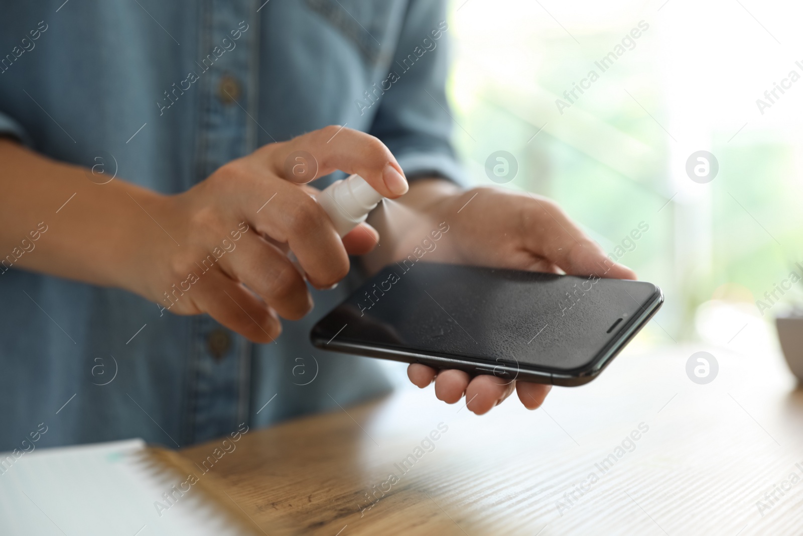 Photo of Woman cleaning smartphone with antiseptic spray at wooden table, closeup