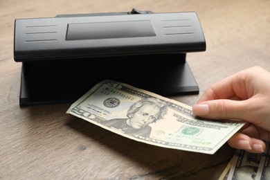 Woman checking dollar banknote with currency detector at wooden table, closeup. Money examination device