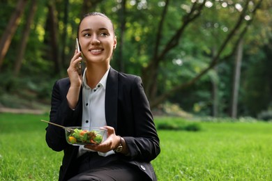 Lunch time. Happy businesswoman with container of salad talking on smartphone on green grass in park, space for text