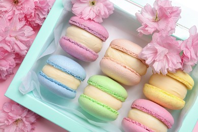 Many delicious colorful macarons in box and flowers on pink background, flat lay