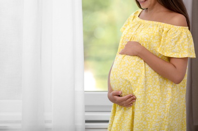 Pregnant woman standing near window at home, closeup