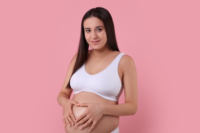 Photo of Beautiful pregnant woman in stylish comfortable underwear making heart with hands on her belly against pink background