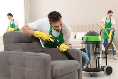 Photo of Professional janitor in uniform vacuuming armchair indoors