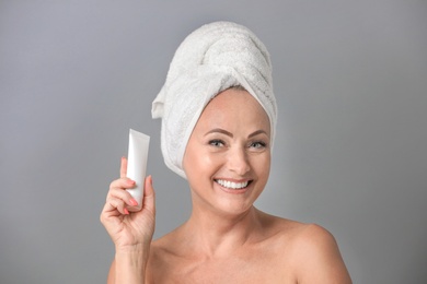 Portrait of beautiful mature woman with perfect skin holding tube of cream on grey background