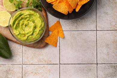 Photo of Bowl of delicious guacamole served with nachos chips and lime on white tiled table, flat lay. Space for text