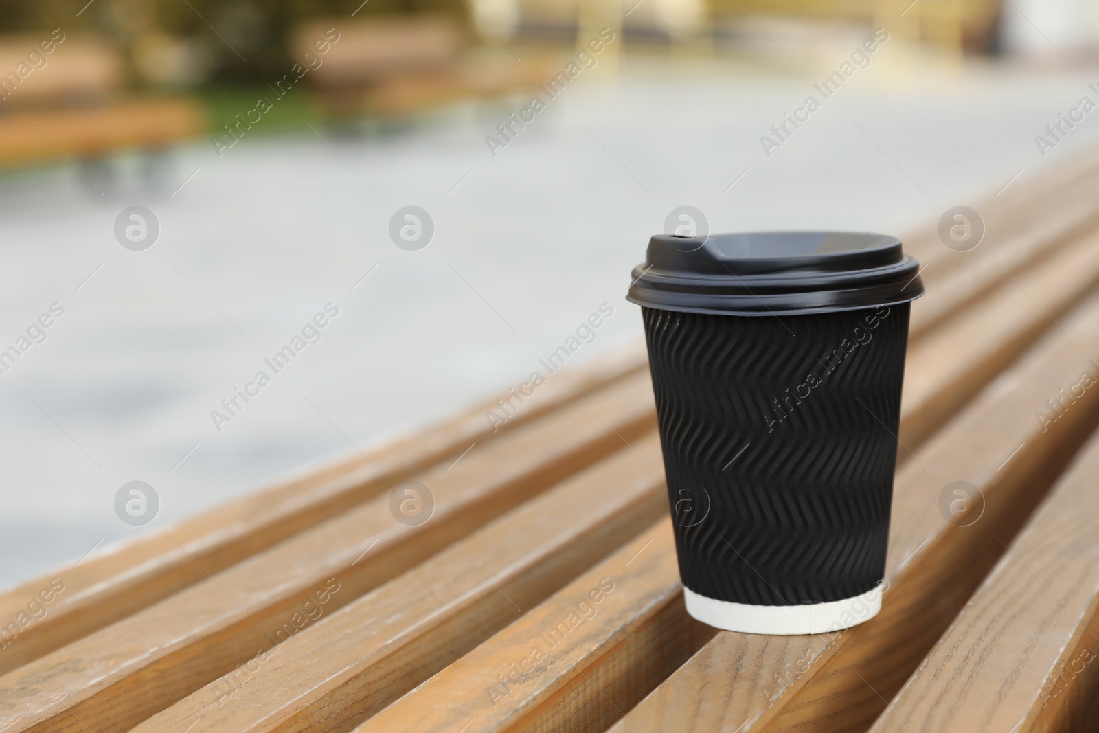 Photo of Black disposable paper cup with plastic lid on wooden bench outdoors. Space for text