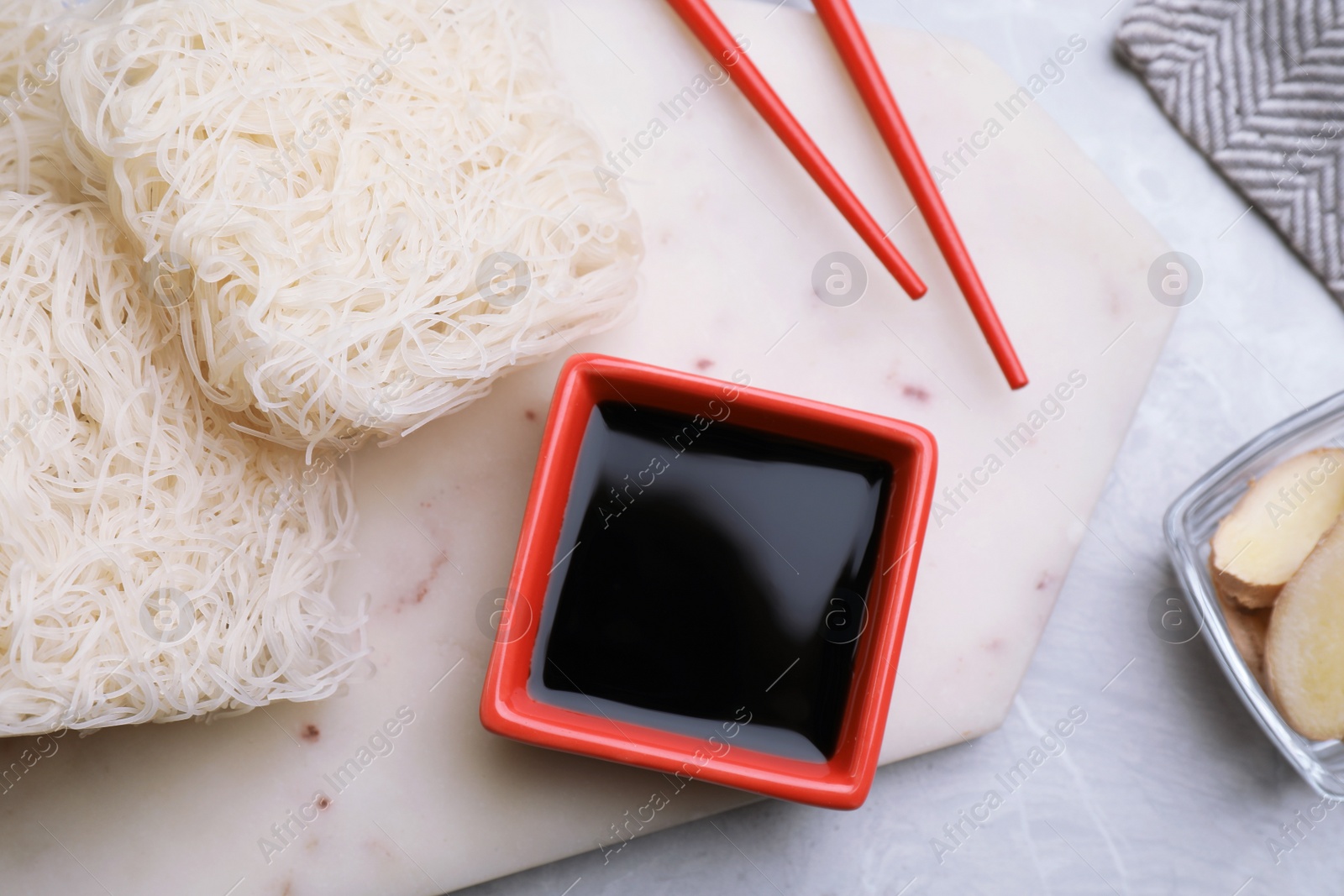 Photo of Uncooked rice noodles, soy sauce and chopsticks on light grey table, flat lay