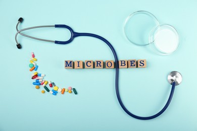 Photo of Word Microbes made with wooden cubes, pills and stethoscope on light blue background, flat lay