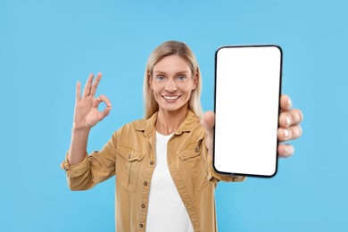 Happy woman holding smartphone with blank screen and showing OK gesture on light blue background