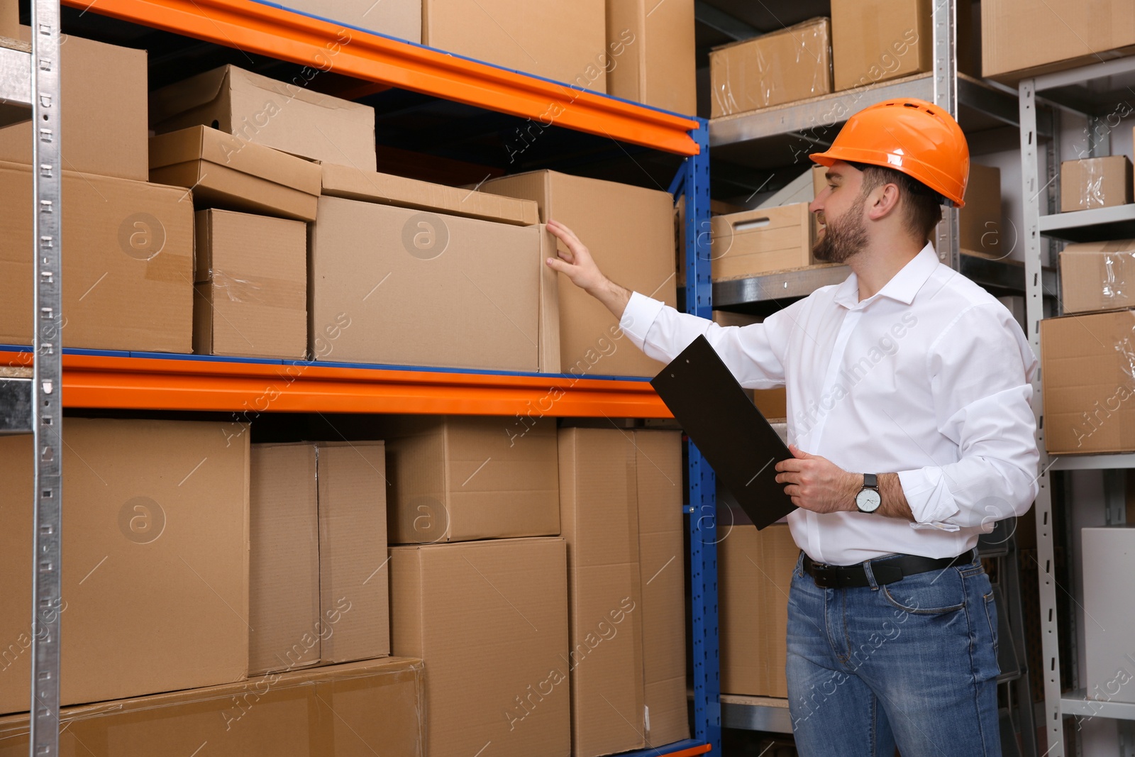 Photo of Young man with clipboard near rack of cardboard boxes at warehouse