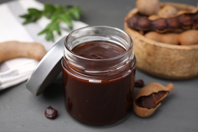 Photo of Jar of tasty tamarind sauce and fresh pods on grey wooden table, closeup