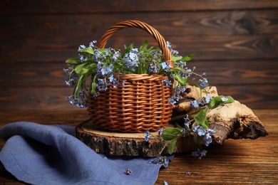 Beautiful forget-me-not flowers in wicker basket, piece of decorative wood and blue cloth on wooden table