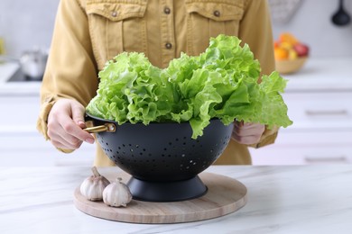 Photo of Woman holding black colander with lettuce at white marble table in kitchen, closeup