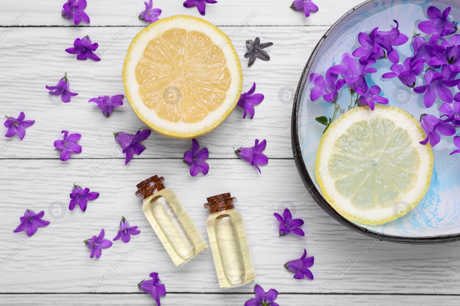 Photo of Bowl with water, lemon, purple flowers and bottles of essential oils on white wooden table, flat lay. Spa composition