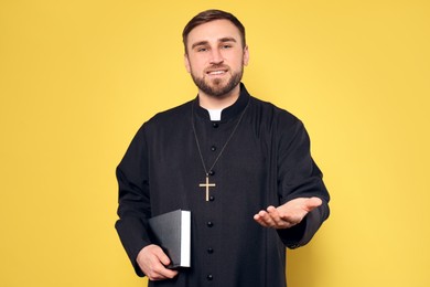 Photo of Priest in cassock with Bible reaching out his hand on yellow background