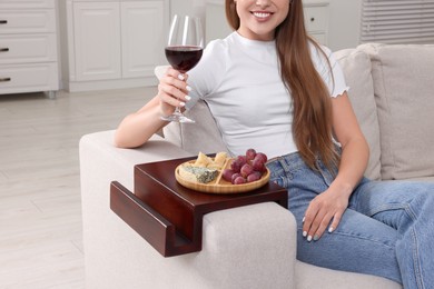 Photo of Grapes and cheese on sofa armrest wooden table. Woman holding glass of wine at home, closeup