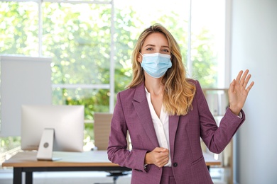 Woman in protective mask showing hello gesture in office. Keeping social distance during coronavirus pandemic